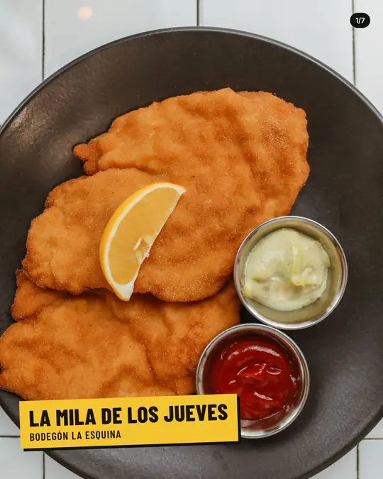 Bodegón La Esquina, lámina 1 de 7: Milanesas doradas en un plato negro sobre azulejos blancos, con una rodaja de limón y potecitos de mayonesa y kétchup. Etiqueta amarilla: La mila de los jueves, Bodegón La Esquina.