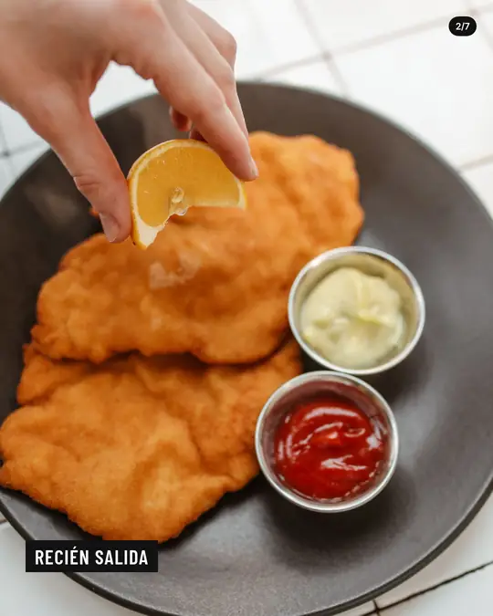 Bodegón La Esquina, lámina 2 de 7: Una mano exprime limón sobre dos milanesas recién hechas, con mayonesa y kétchup al costado.