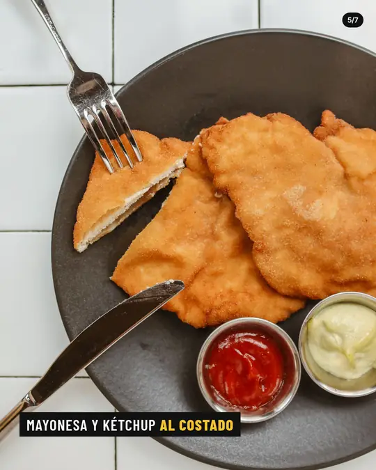 Bodegón La Esquina, lámina 5 de 7: Plato de milanesas con un pedazo cortado, tenedor, cuchillo y potecitos de mayonesa y kétchup.