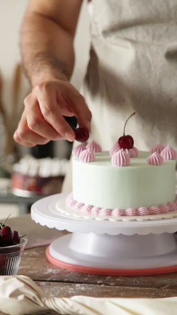 Manos colocan cerezas sobre una torta verde agua decorada con merenguitos rosas